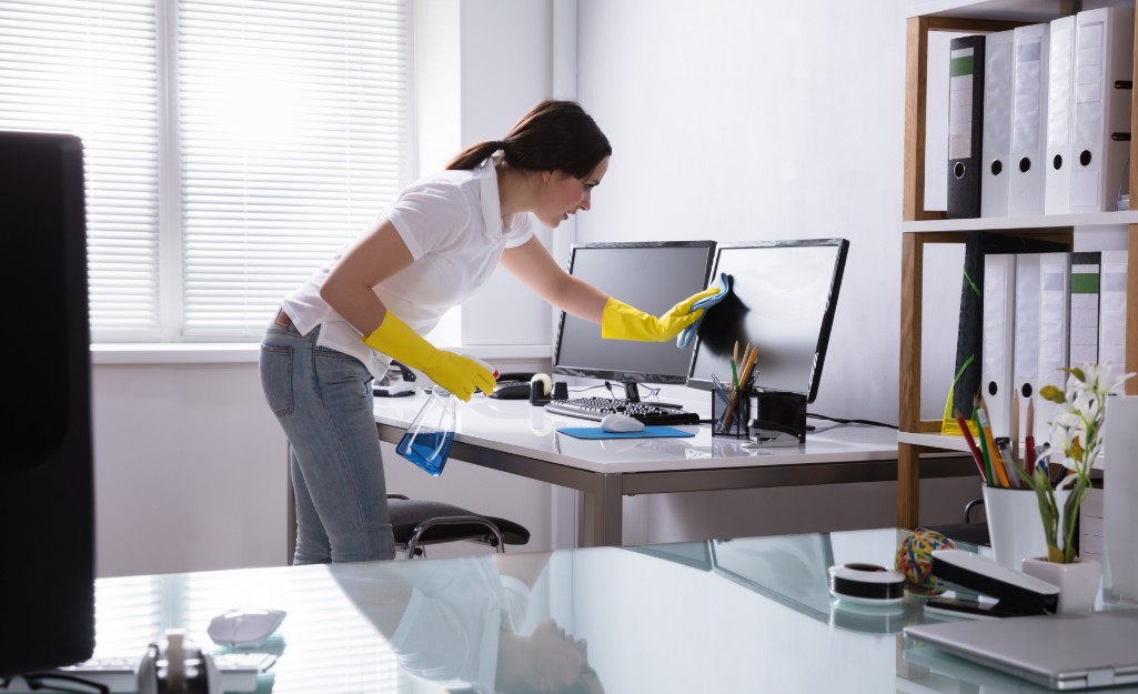 Cleaner dusting a computer screen at a desk in a bright office